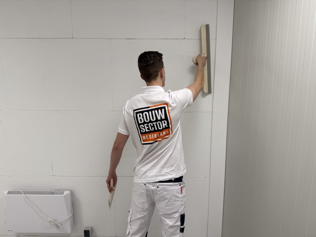 Stukadoor met het logo van Bouwsector Nederland op zijn rug werkt met de spaan hoog op een wand van gipsblokken