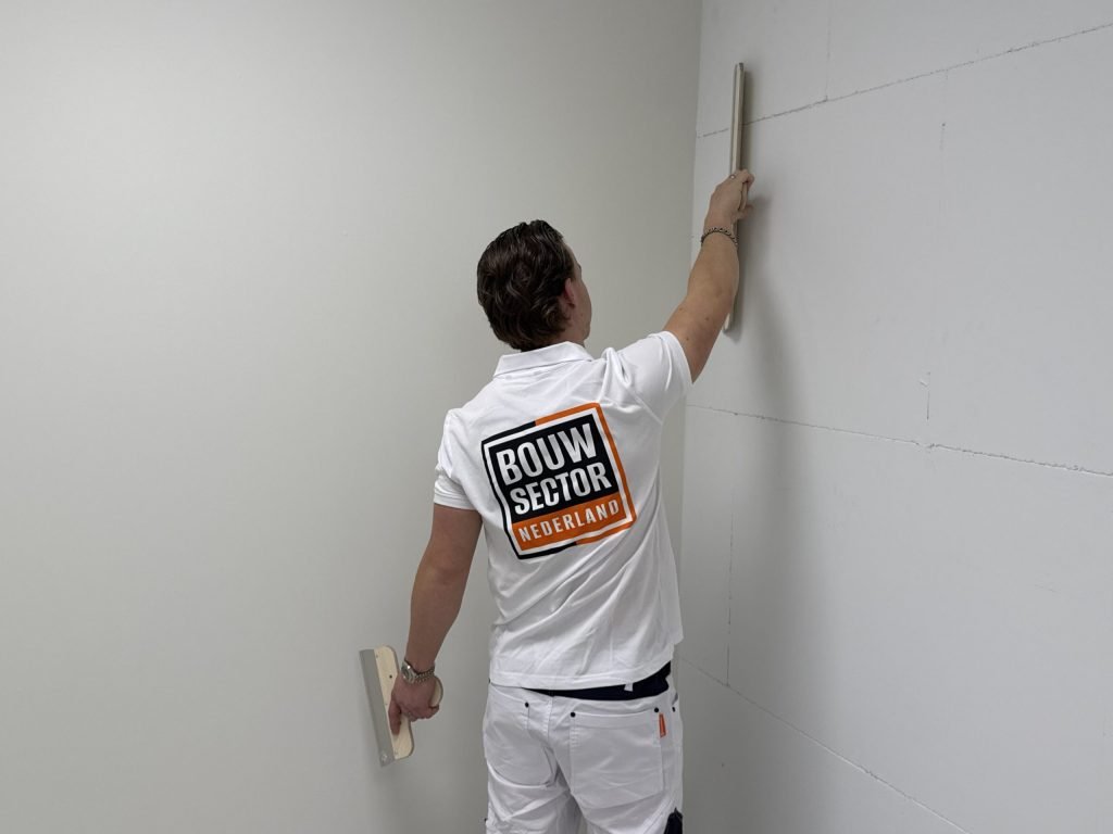 Stukadoor in wit werkpak trekt met een lange spaan een wand in de hoek van een kamer vlak