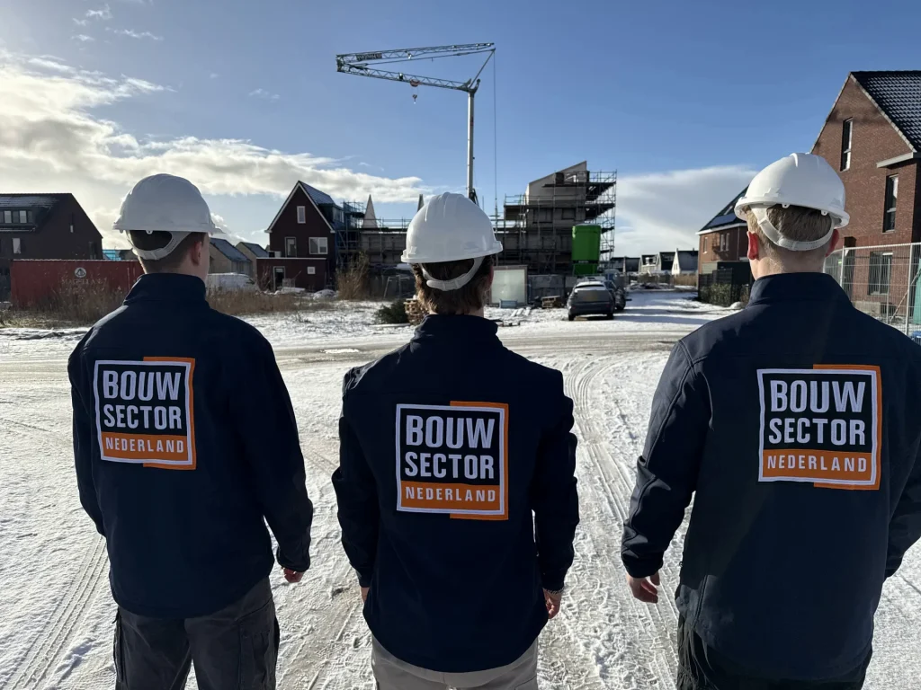 Drie vakmensen van Bouwsector Nederland met witte helm en donkerblauwe jas kijken naar een nieuwbouwwijk met een bouwkraan