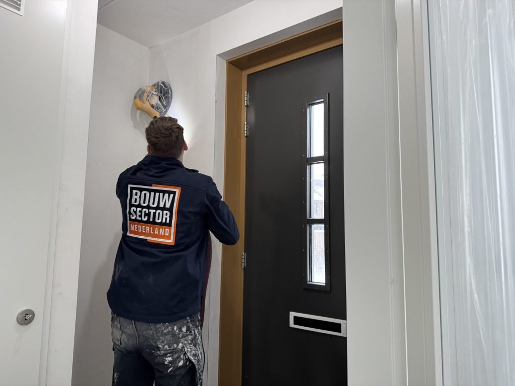 Vakman van Bouwsector Nederland schuurt met een wandschuurmachine de muur boven een voordeur