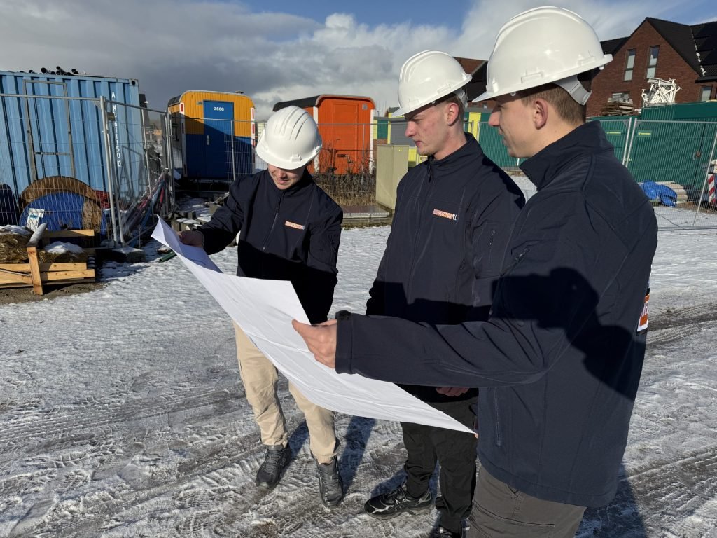 Drie vakmensen van Bouwsector Nederland met witte helm bekijken een bouwtekening op een besneeuwd nieuwbouwterrein